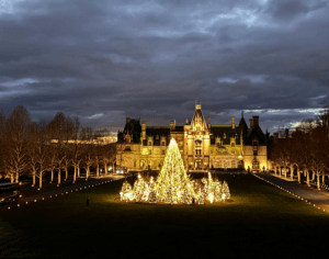 A beautifully illuminated mansion with a large Christmas tree in the foreground, set against a dramatic evening sky.