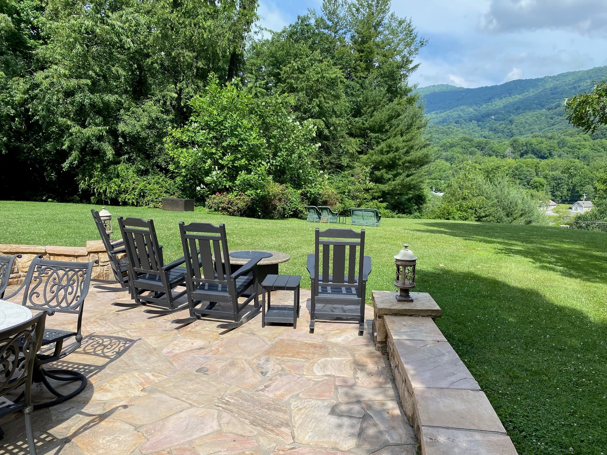A stone patio with black rocking chairs overlooks a lush green landscape and distant mountains under a partly cloudy sky. A stone patio with black rocking chairs overlooks a lush green landscape and distant mountains under a partly cloudy sky.