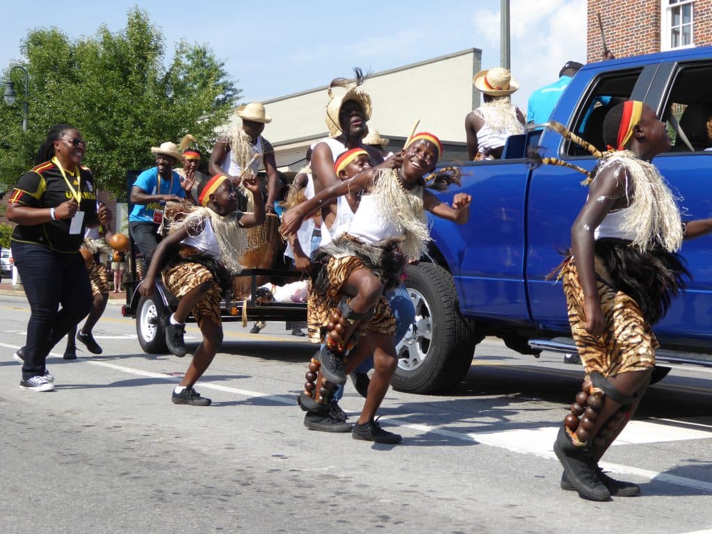 A group of performers in traditional attire dance energetically alongside a blue truck during a parade.