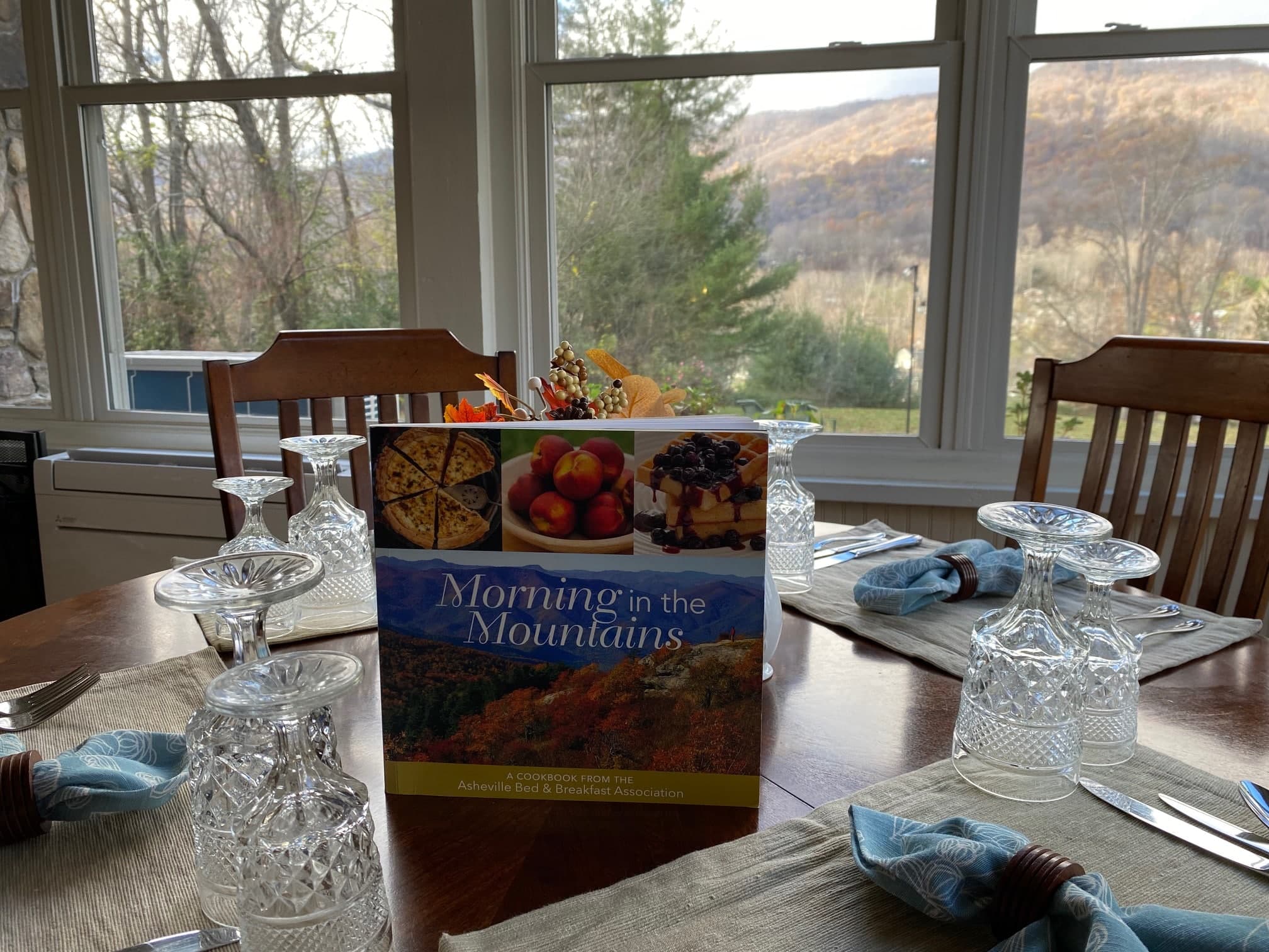 A dining table set with crystal glassware and a cookbook titled "Morning in the Mountains," with a scenic view of mountains and trees in the background. A dining table set with crystal glassware and a cookbook titled "Morning in the Mountains," with a scenic view of mountains and trees in the background.