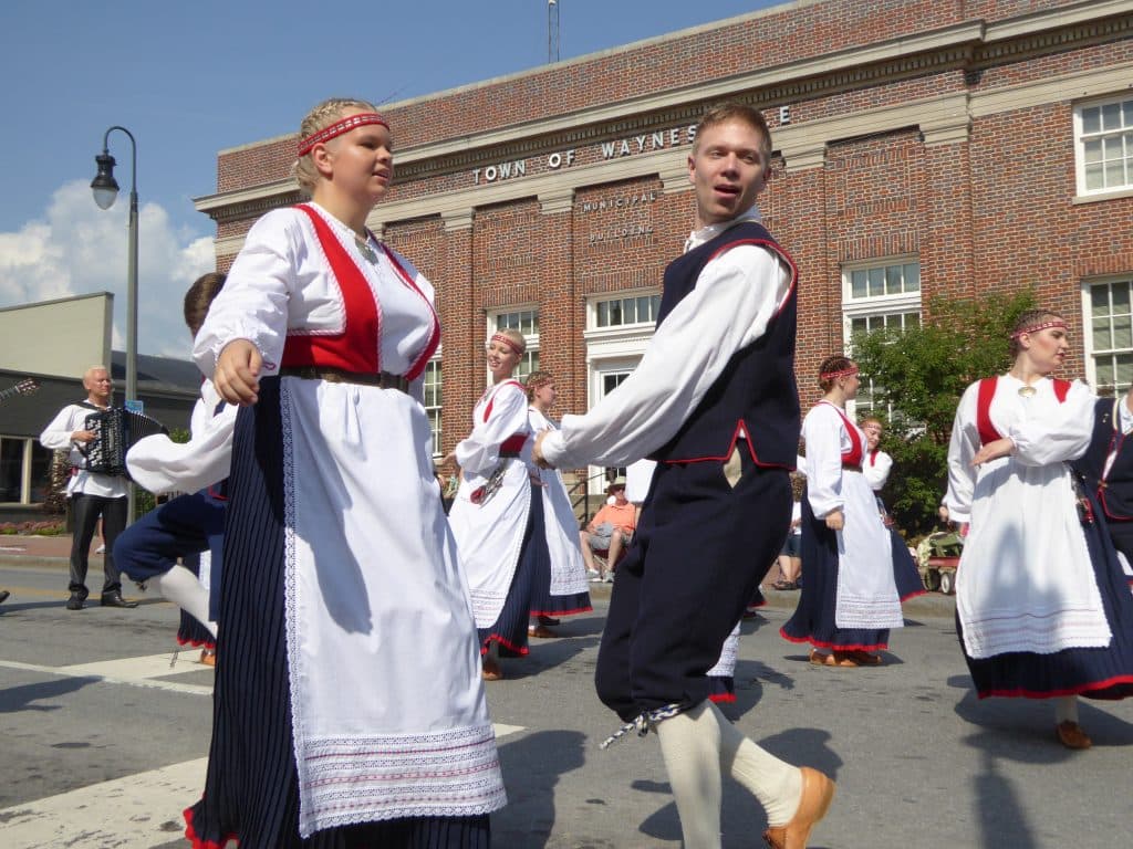 Dancers in traditional costumes perform in front of a brick municipal building during a lively parade.