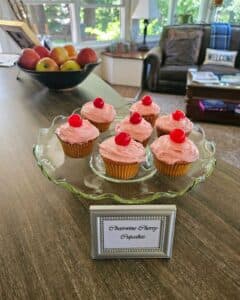 A glass cake stand with six frosted cupcakes topped with cherries, alongside a sign that reads "Strawberry Cherry Cupcakes."