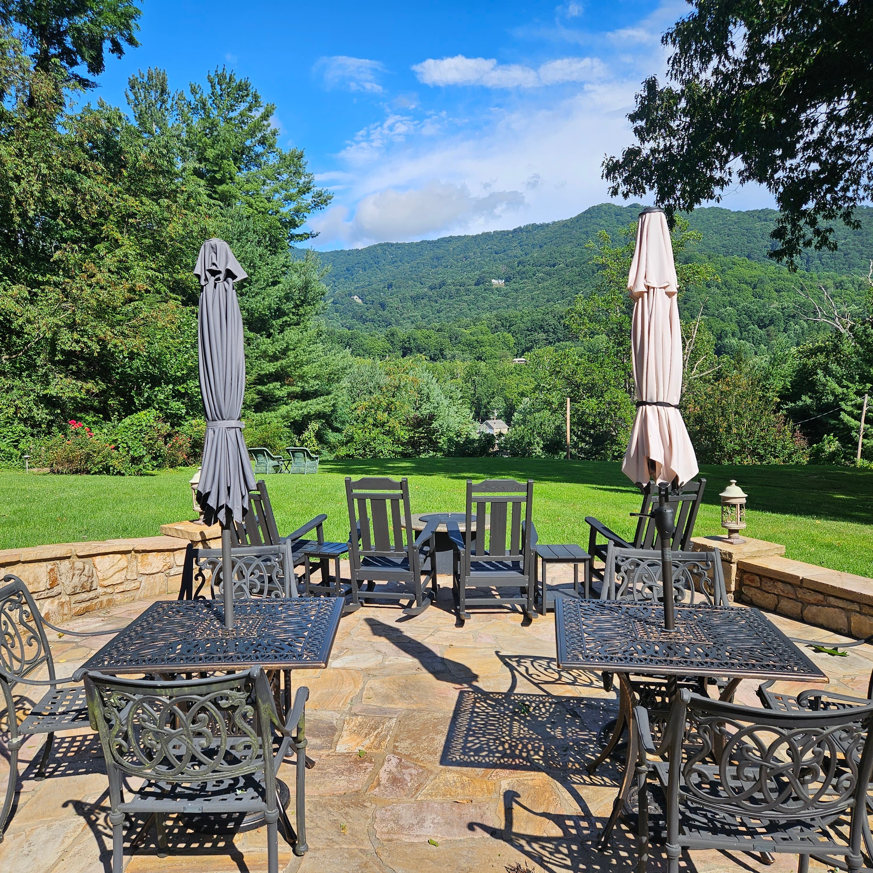 A patio with metal tables and chairs overlooks a lush green landscape and mountains under a blue sky.