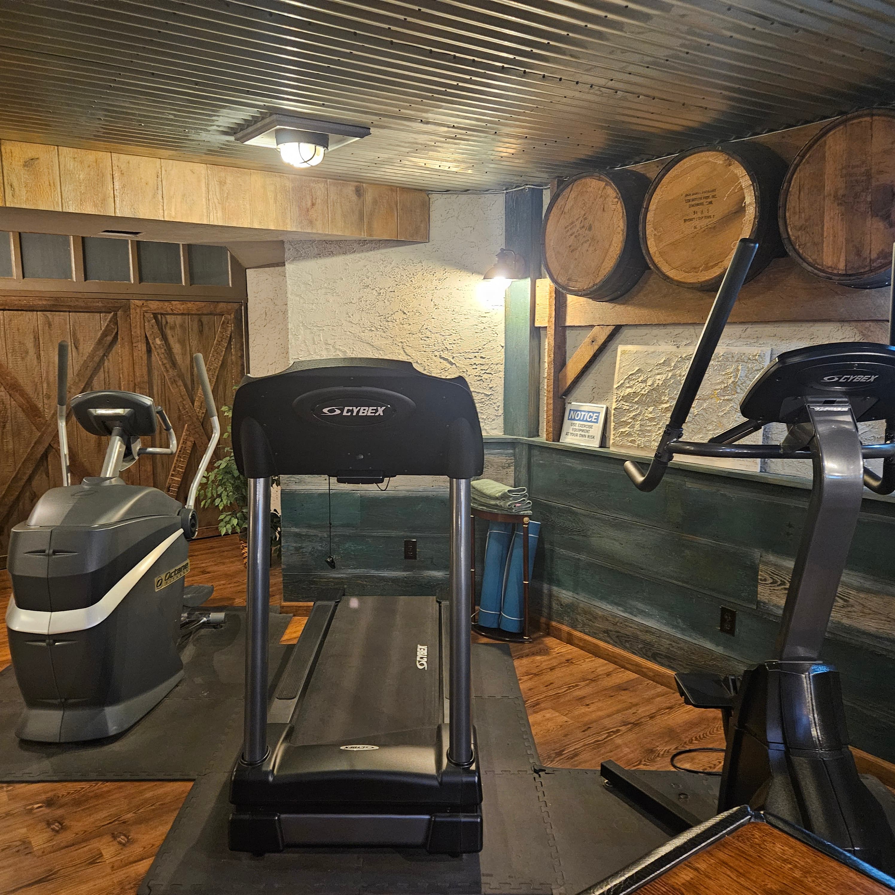 A small home gym featuring a treadmill, an elliptical machine, and wooden barrels on the wall.