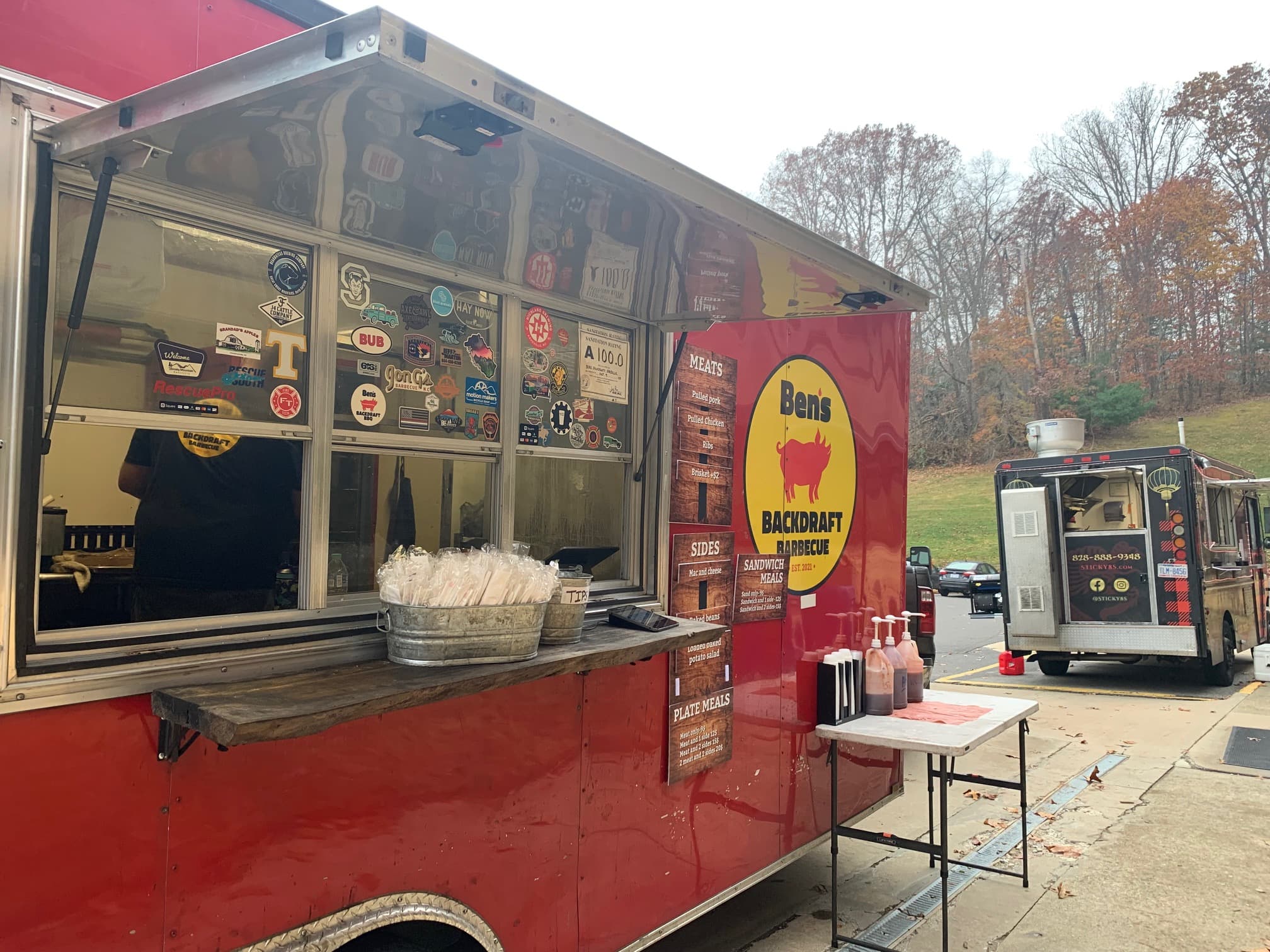 A red food truck named "Ben's Backdraft Barbecue" displays a menu and various stickers, with another truck visible in the background.