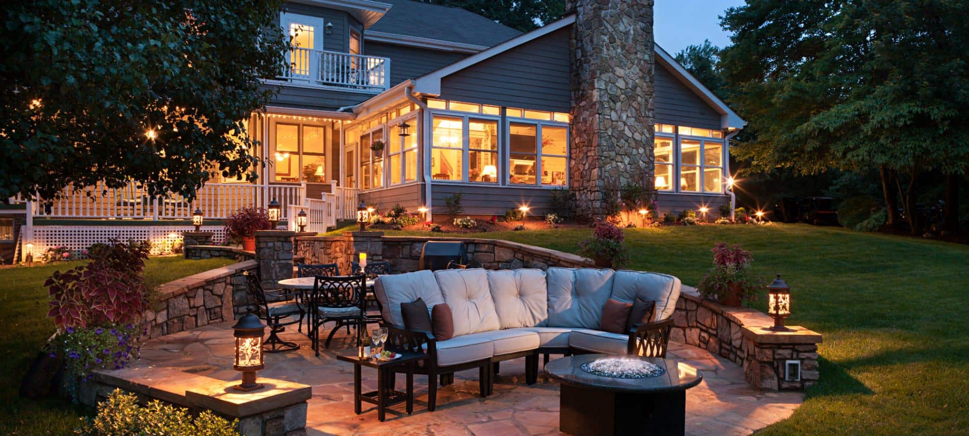 A cozy outdoor patio featuring a sectional sofa, fire pit, and illuminated garden in the evening.