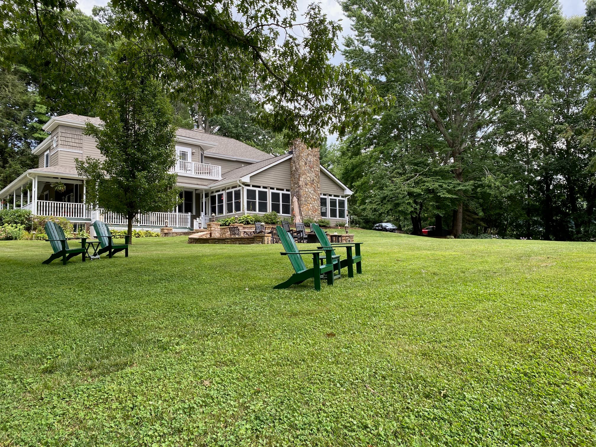 A grassy yard featuring green adirondack chairs in front of a large house surrounded by trees.
