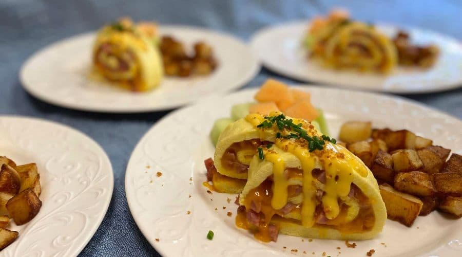 A plate of rolled omelette topped with cheese and accompanied by potato cubes and fruit.