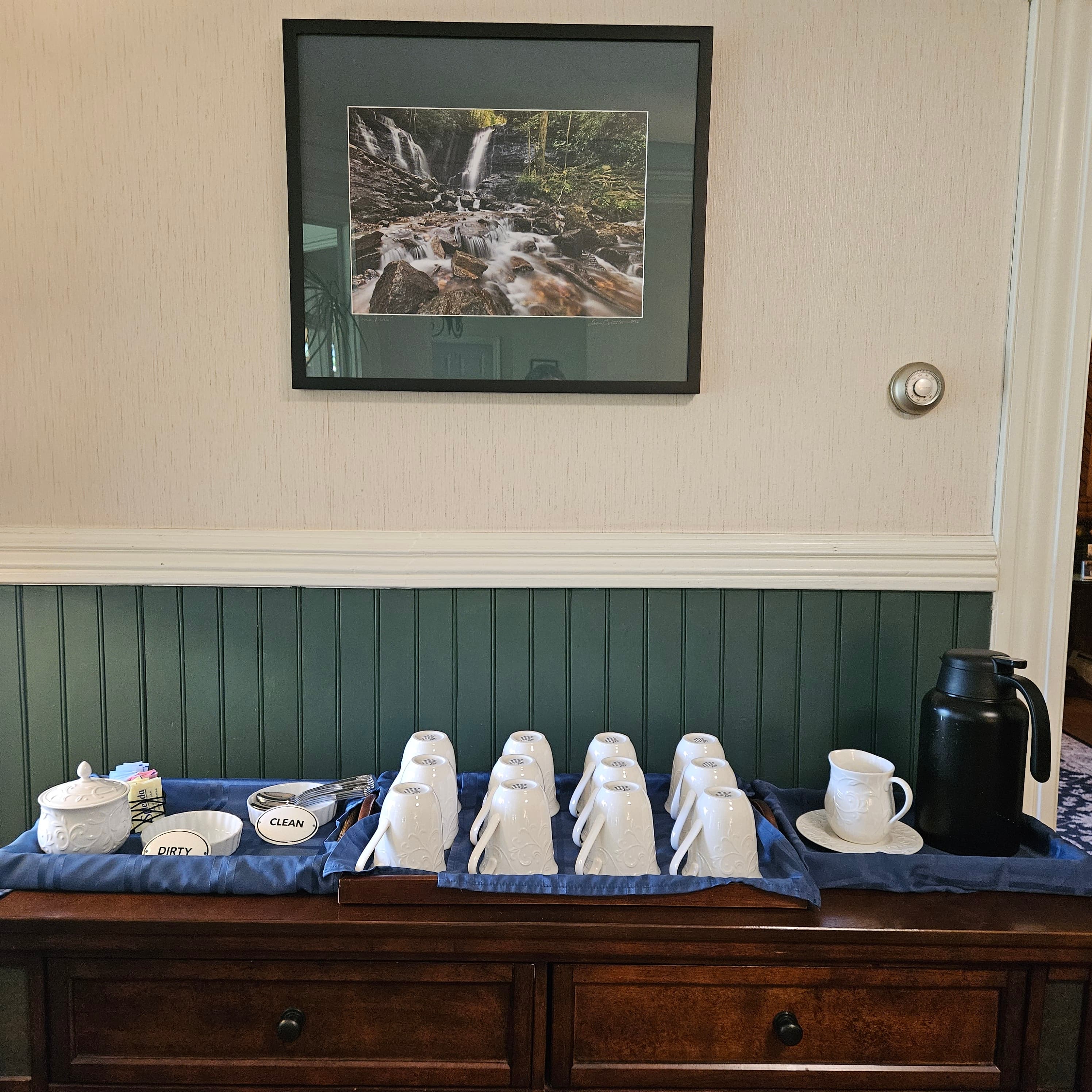 A wooden table holds clean and dirty cups, a coffee pot, and a decorative photograph of a waterfall above.
