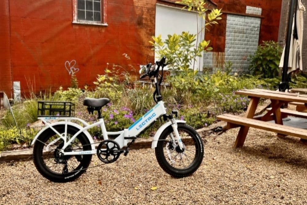A white electric bike is parked on gravel near a wooden picnic table in a green, overgrown garden.