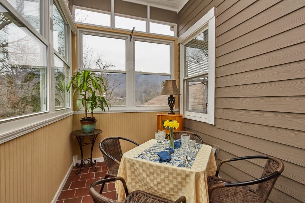 A cozy sunroom with a small table set for two, featuring a lamp, potted plant, and scenic views through large windows.
