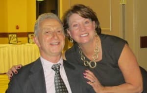 A smiling man sits in a chair while a woman stands beside him, both enjoying a moment together.