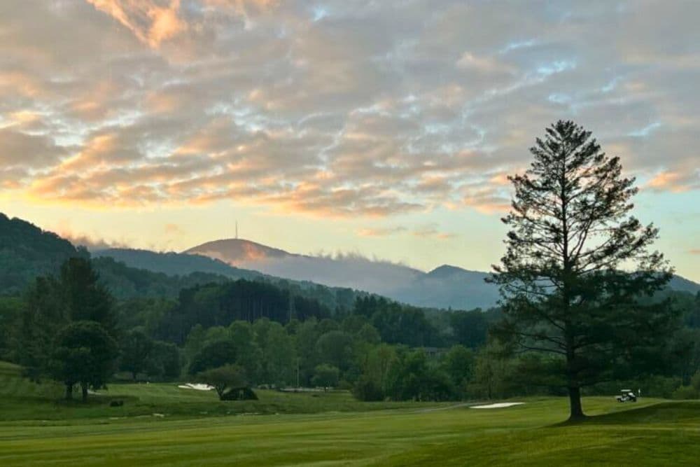 A serene landscape featuring a golf course, rolling hills, and a mountain under a colorful sky at sunrise.