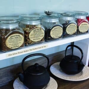 A shelf displaying labeled jars of tea ingredients above two traditional black teapots.
