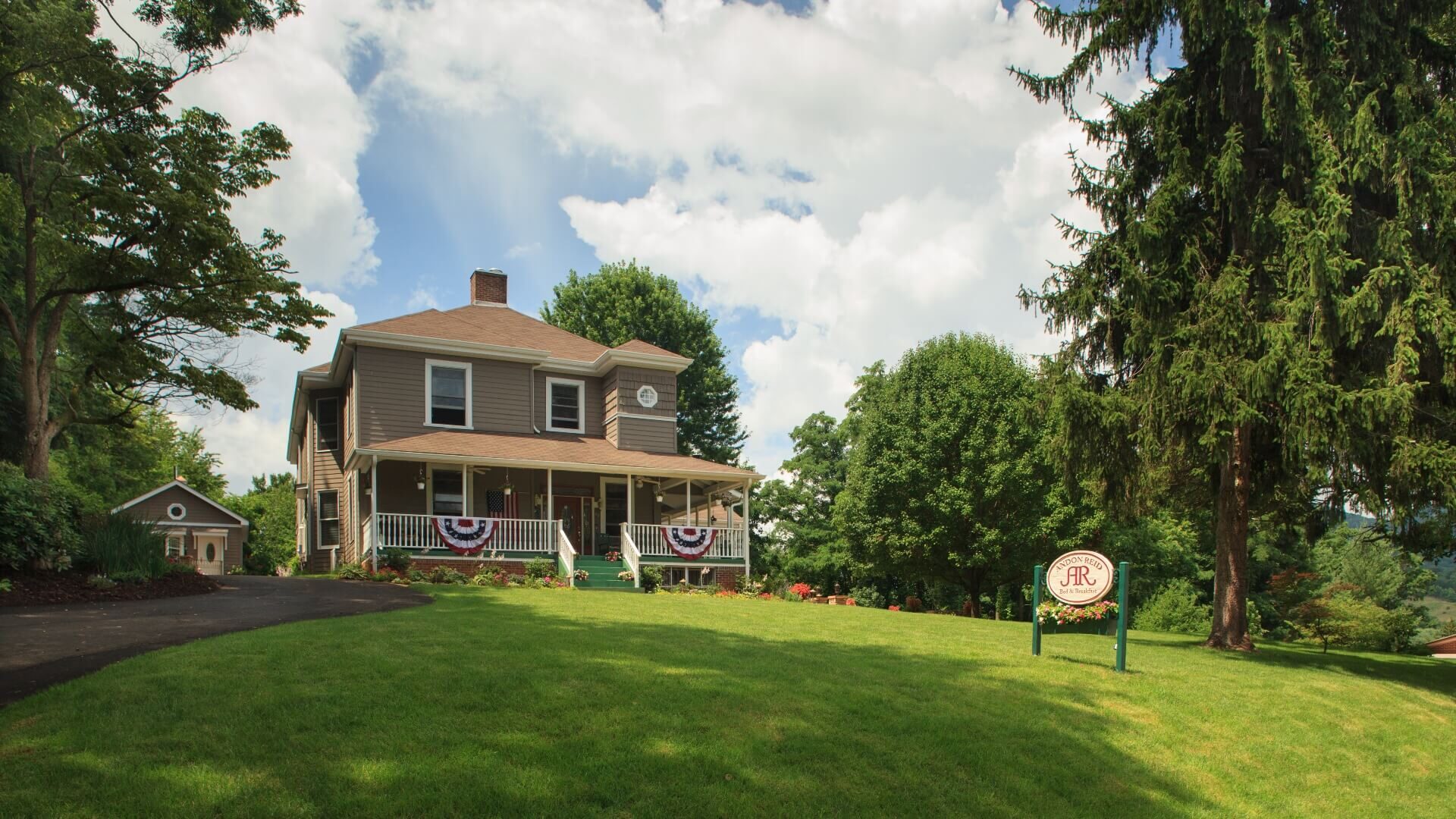 A stately two-story home surrounded by lawn and large green trees with blue skies above and sign on the front yard