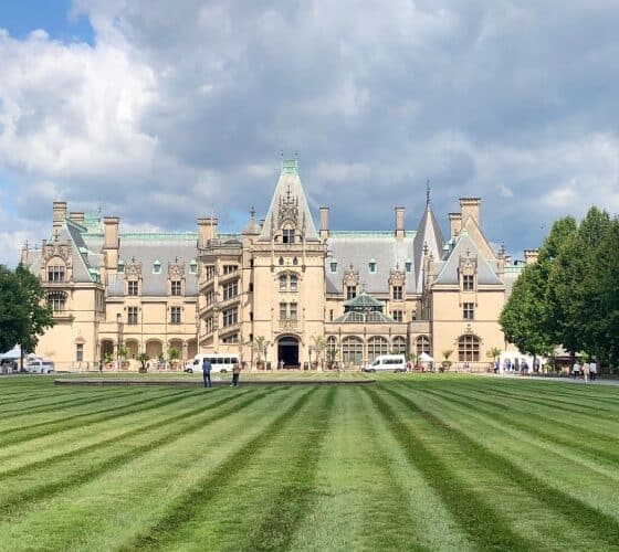Exterior view of the front of an expansive castle-like estate surrounded by pristinely cut grass and blue skies above