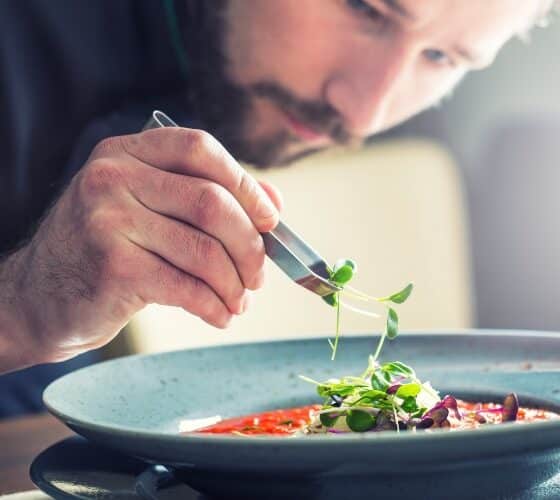 A chef holding tongs placing some leafy greens on a round plate of pasta