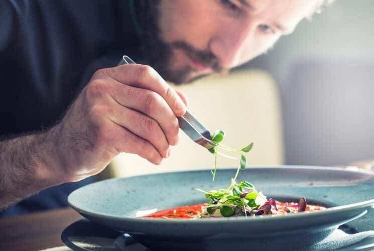 A chef holding tongs placing some leafy greens on a round plate of pasta