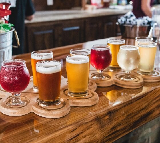 A wood counter at a bar with a flight of 10 different glasses of beer of various colors