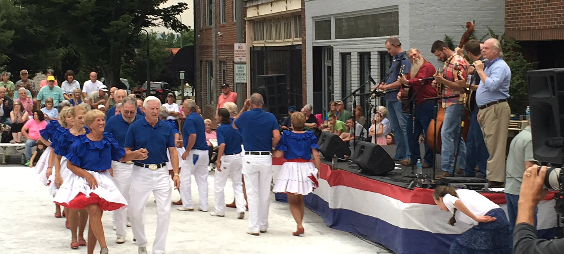 City celebration, band playing on a raised platform, couples in blue shirts and white slacks and skirts dancing