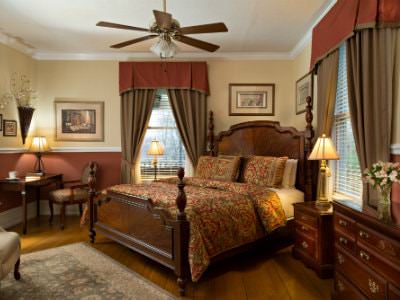 Beige guest room with ceiling fan, wood four poster bed, elegant furnishings, wood floors, and two windows
