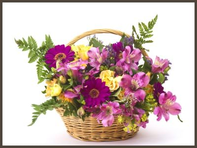 Tan wicker flower basket with handle filled with pink, violet and yellow flower with green leafy stems