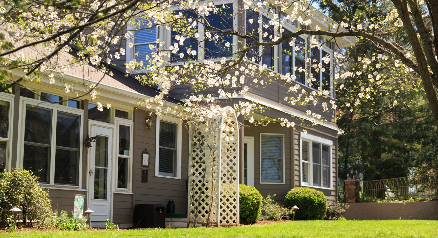 Outdoor view of a large two-story home with many windows, green lawn with shrubs and trees with white flowers