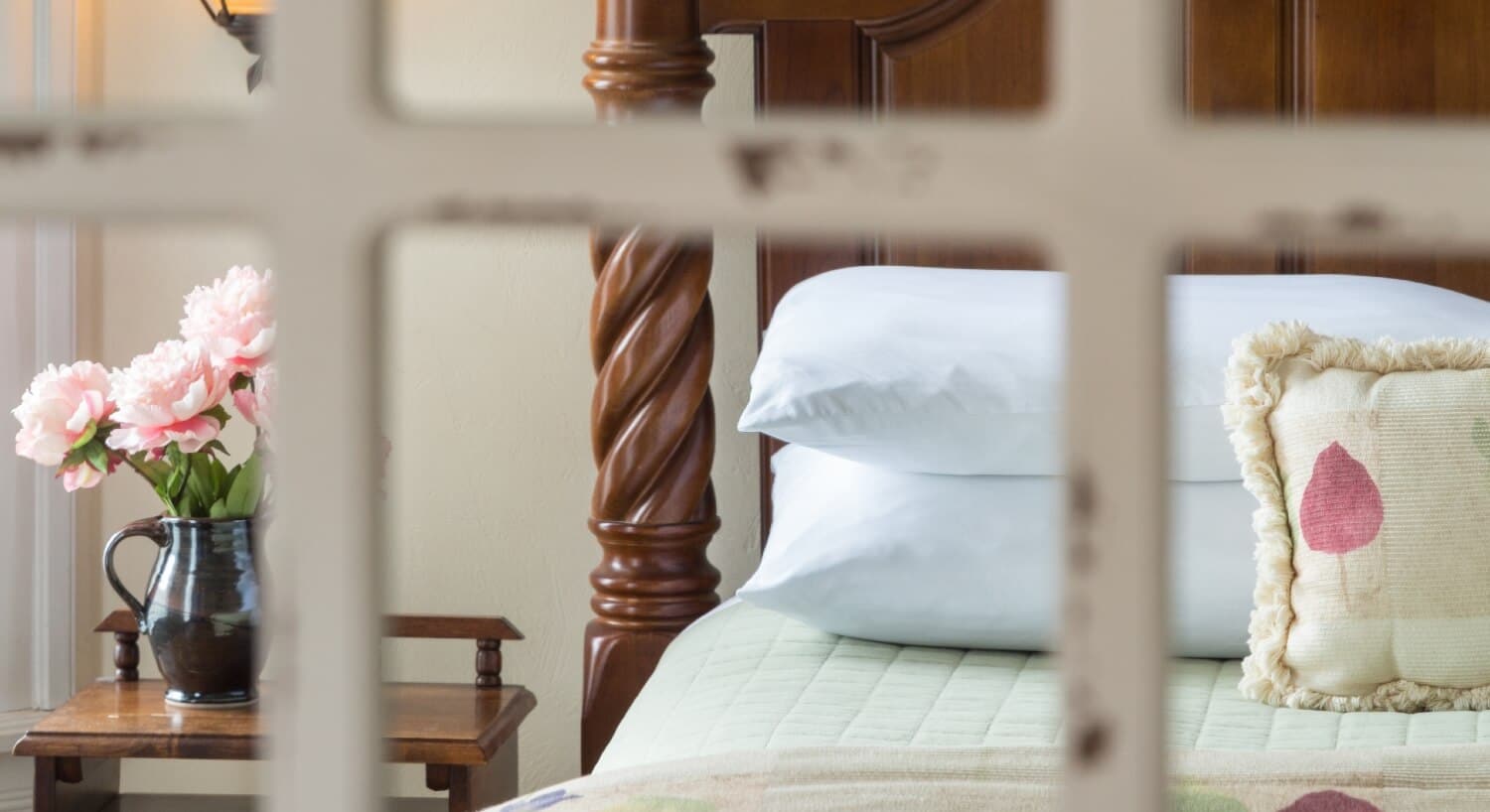 Upclose view through a framed window of a bed with two stacked white pillows and a side table wth a vase of pink flowers