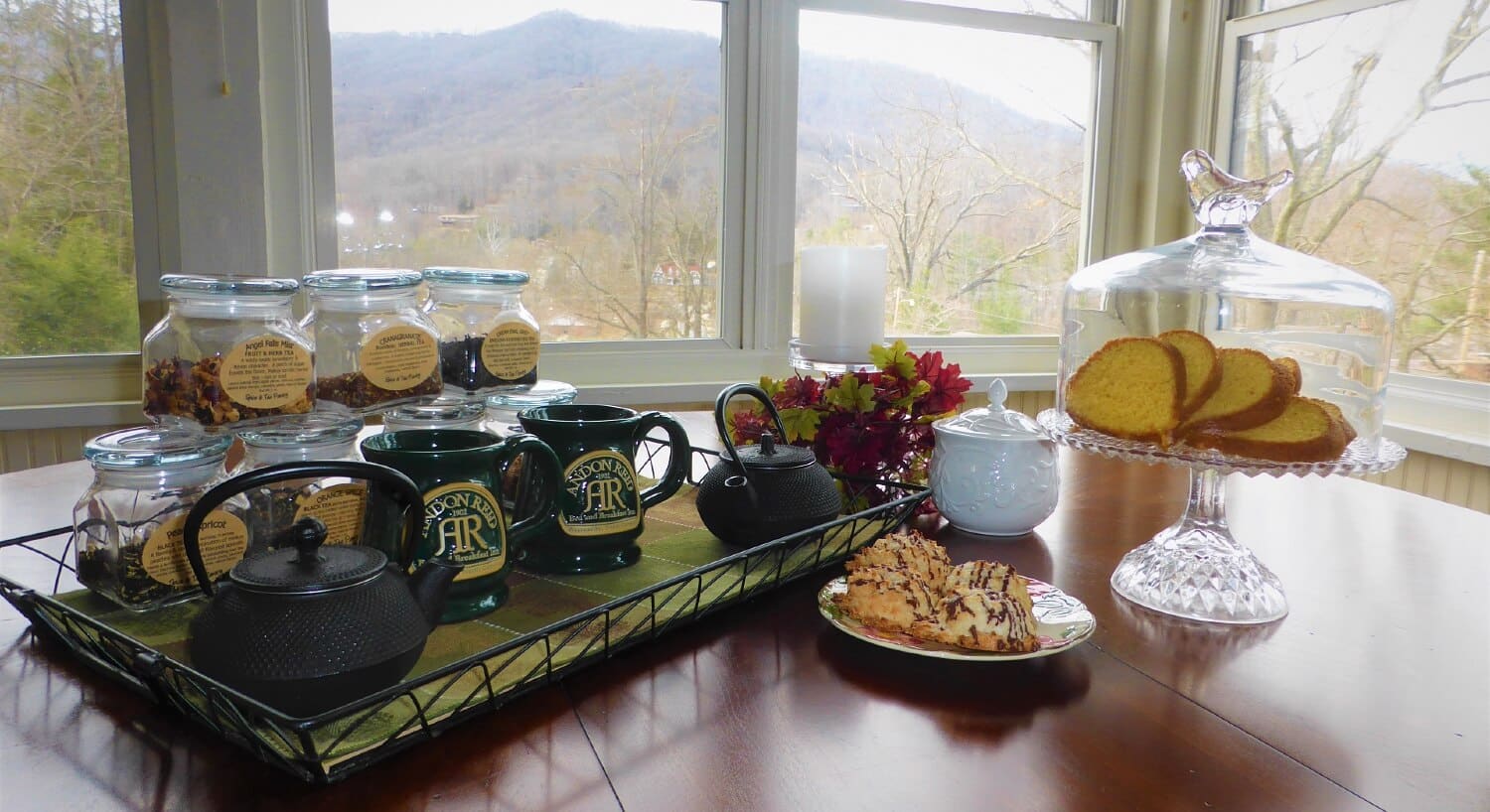 Dining table with tray of mugs and two teapots, jars of nuts and glass cake stand with slices of cake