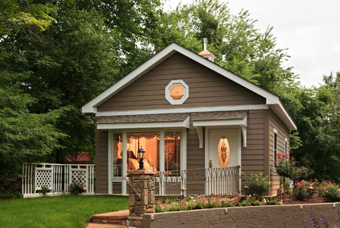 Brown sided cabin with white trim, gable roof, surrounded by flowers, green grass and trees