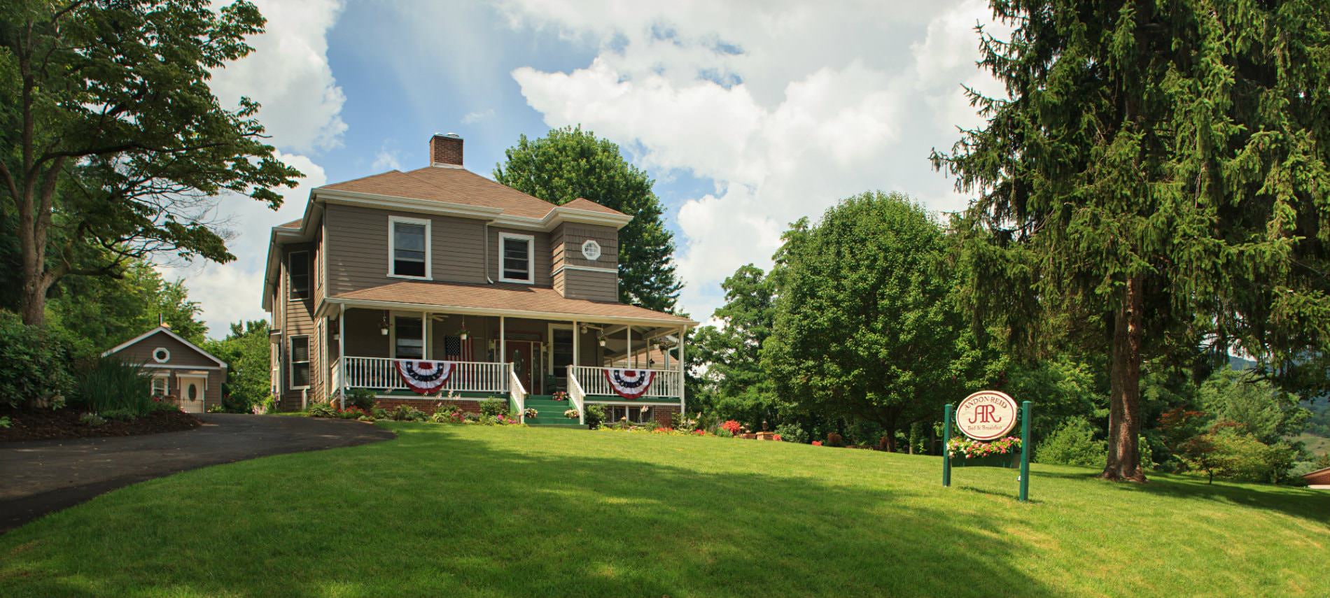 Brown sided two story home with white trim, hip roof, surrounded by green grass, lush green trees and blue skies