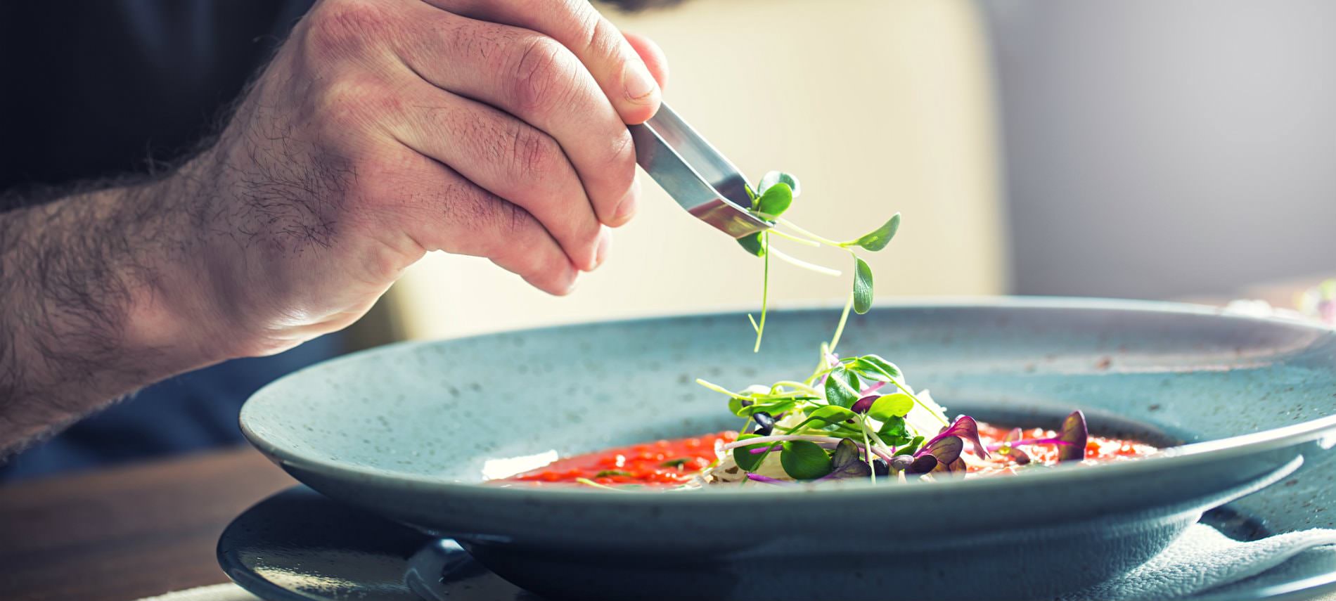 Man's hand holding silver tongs placing fresh herbs on homemade soup in a turquiose bowl