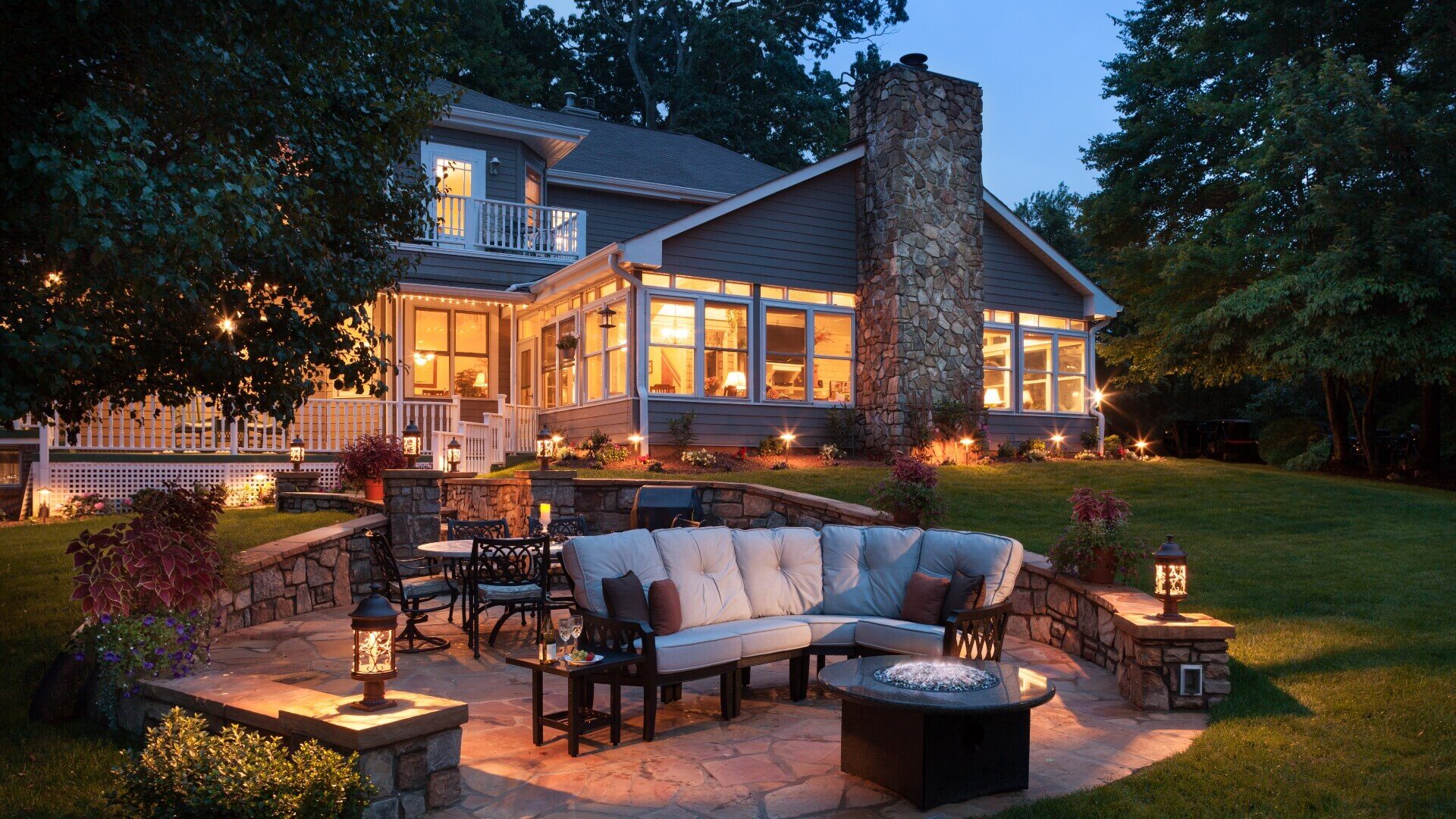 Backyard area of a large home lit up at night with stone patio with table and chairs and couch facing a fire pit