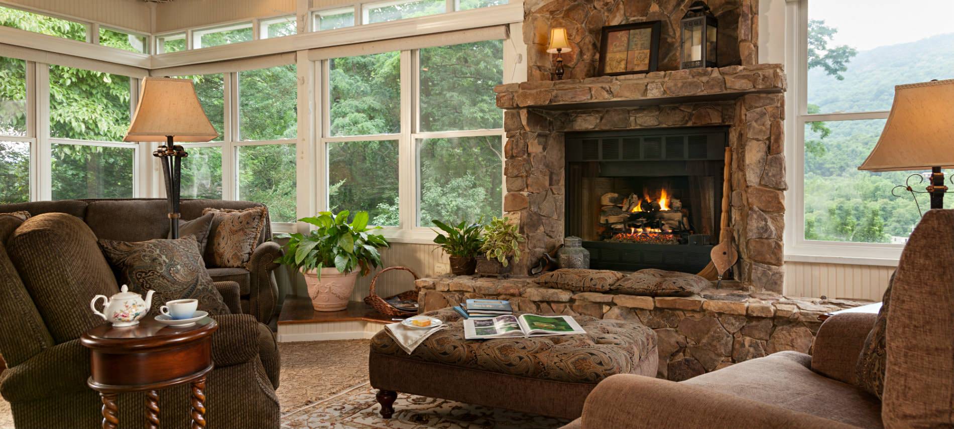 Beautiful sun room surrounded with windows, large stone hearth and fireplace, upholstered furniture and lush green views