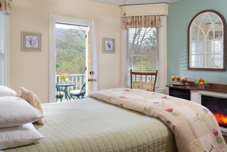 Cozy bedroom in hues of cream and sage with electric fireplace, rocking chair and doorway open to an outdoor patio overlooking the mountains