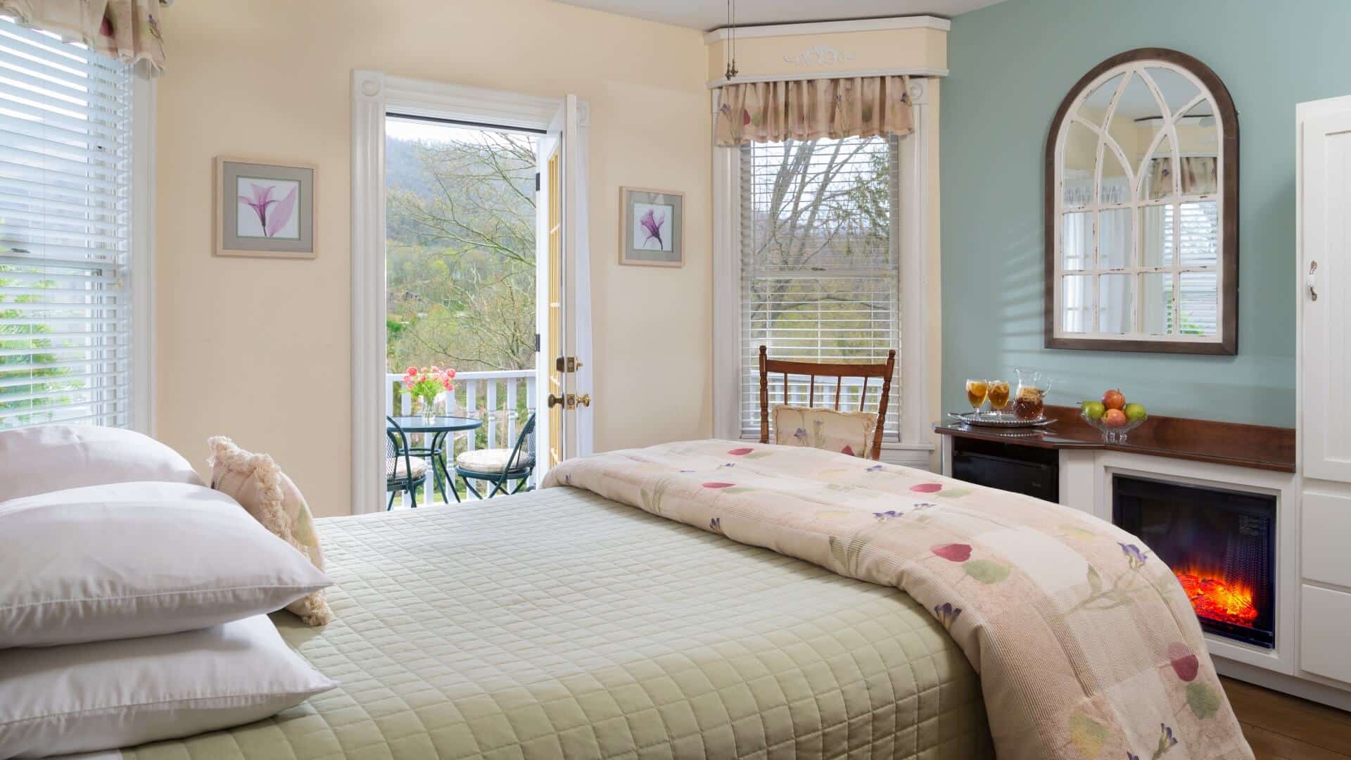 Cozy bedroom in hues of cream and sage with electric fireplace, rocking chair and doorway open to an outdoor patio overlooking the mountains