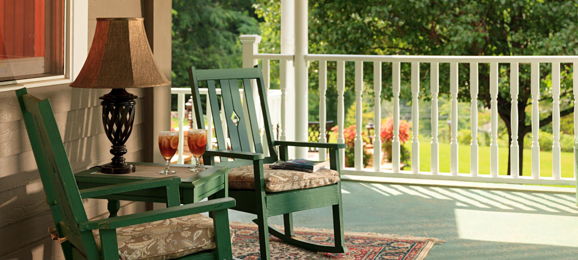 Covered porch with white railing, two green rocking chairs with cushions, table with lamp and view of green landscaping