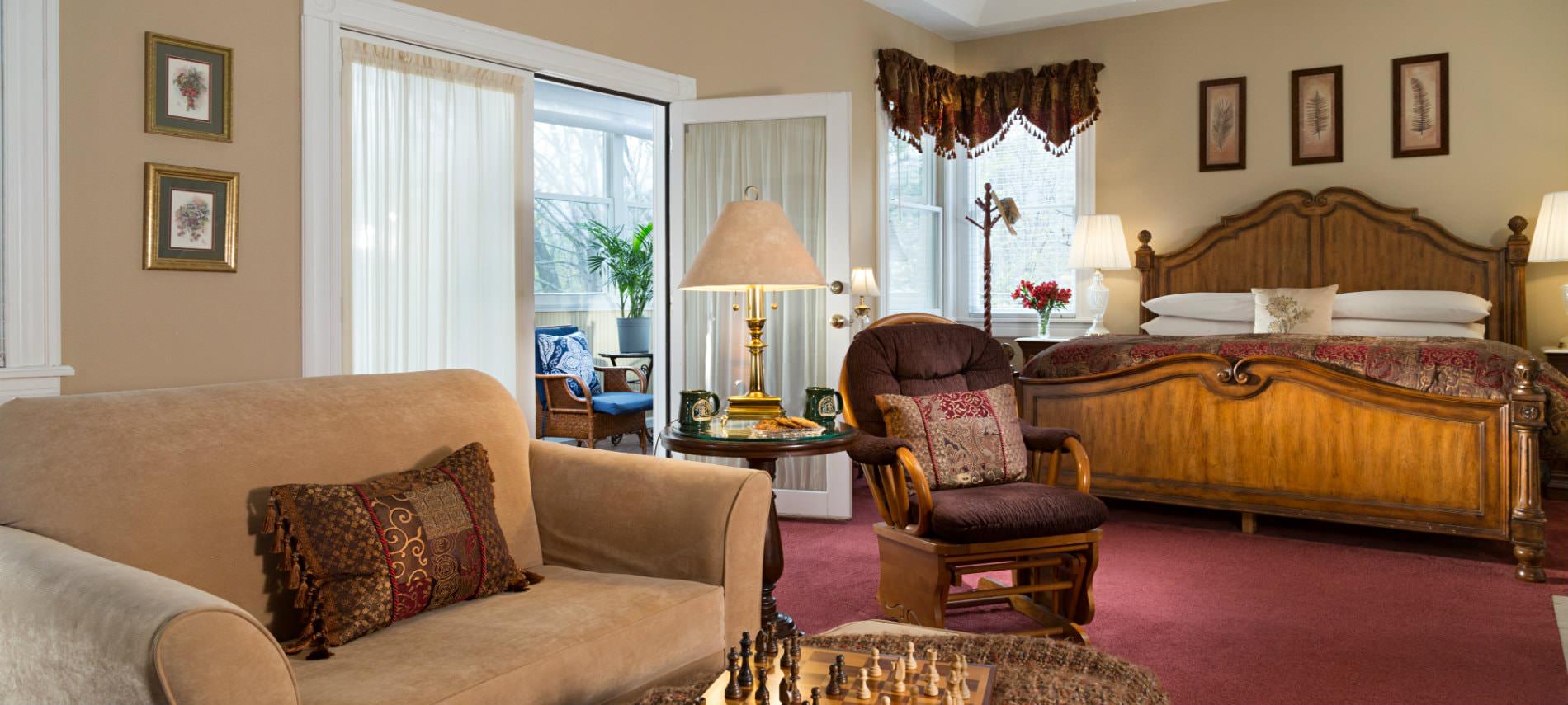 Spacious beige guest room, red carpeting, large wood bed, sitting area, natural light, French door to a cheery sunroom