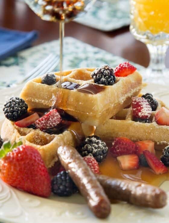 Breakfast plate of waffles, berries and two sausage links with a glass of orange juice