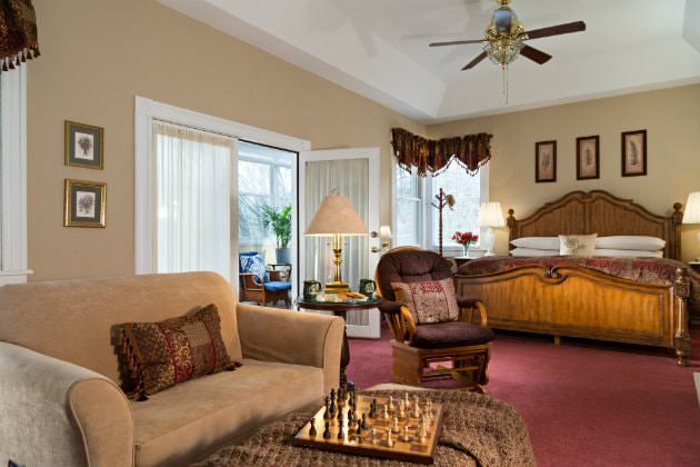 Spacious beige guest room, red carpeting, large wood bed, sitting area, natural light, French door to a cheery sunroom
