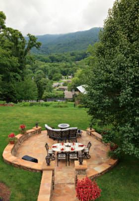 Peachy tan stone walkway and sunken patio with tables and chairs surrounded by flowers, green gress and trees