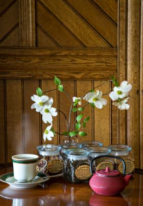 Table topped with fresh white flowers with green leafy stems, jars of loose leaf tea, teacup and saucer, and red teapot