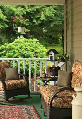 Covered porch with white railing, wicker furniture with colorful cushions, and a view of lush green landscaping