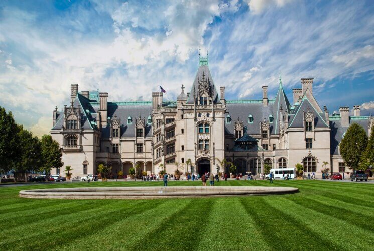 Exterior view of the front of an expansive castle-like estate surrounded by pristinely cut grass and blue skies above