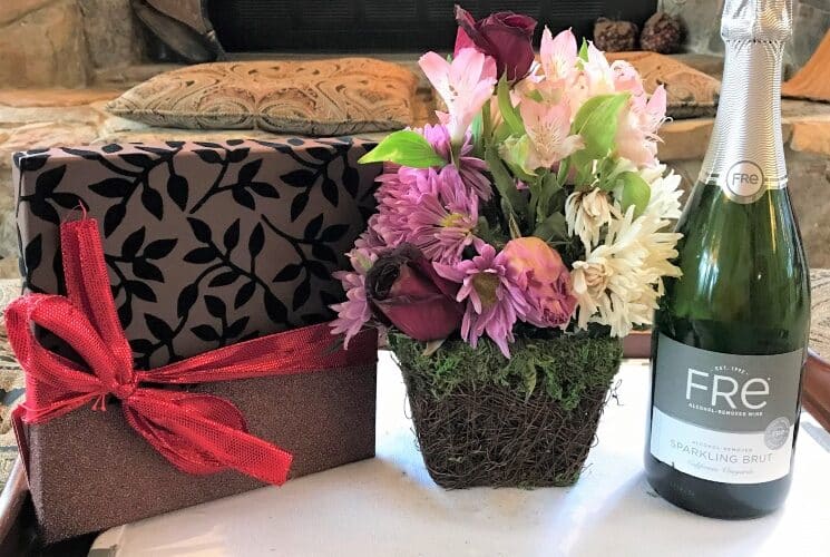 Table holding bottle of champagne, basket of flowers and box of chocolates with a red bow