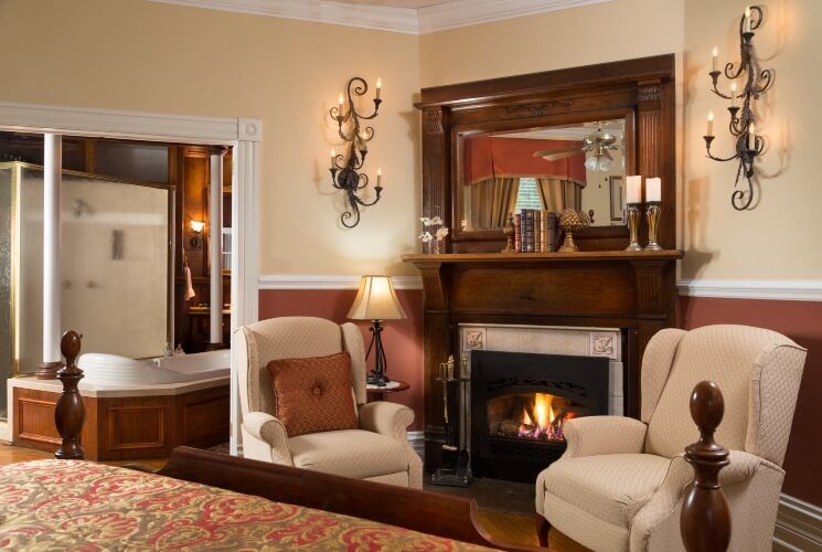 Sitting area in a bedroom with two cream wingback chairs in front of a fireplace, near a doorway open to a bathroom
