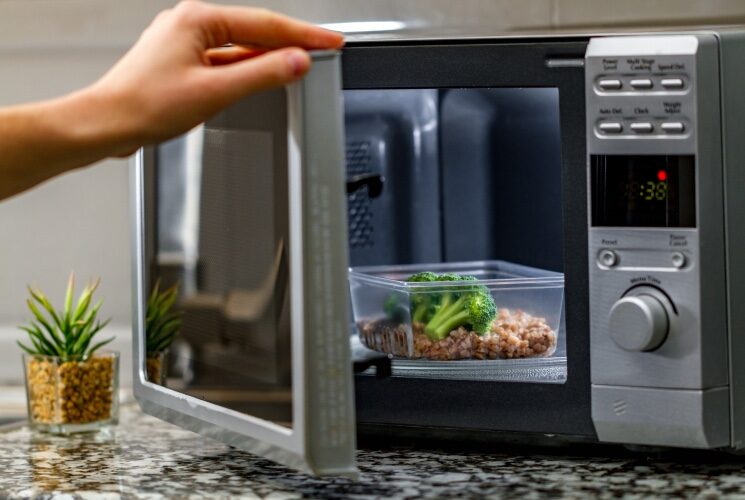 A person holding a microwave door open with a container of food inside
